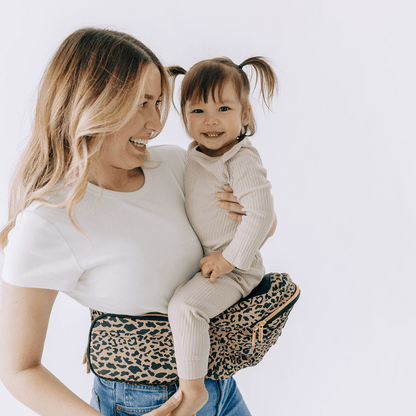 Tushbaby hip carrier for babies and toddlers, with adjustable straps and padded support, shown in a natural beige color.